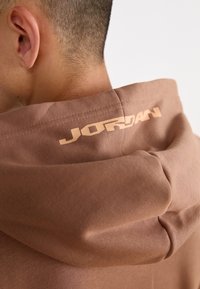 Sweat à capuche marron avec une texture douce, arborant un grand logo "JORDAN" beige au niveau de la nuque à l'arrière. Design simple, sans motifs supplémentaires.