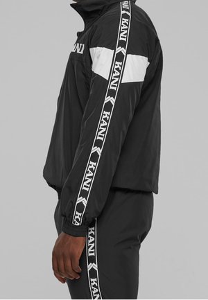 Personne portant un survêtement noir avec des bandes diagonales blanches et des bandes avec le logo "KANI" sur la manche et la jambe, debout de côté.
