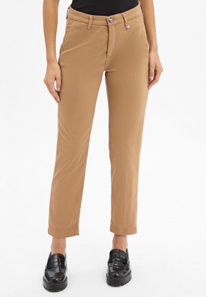 Woman wearing tan straight-leg pants and black loafers, standing against a plain white background.