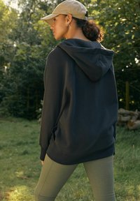 Tmavá mikina s veľkou kapucňou, v kombinácii s olivovozelenými legínami. Modelka má na sebe béžovú čiapku, ktorá vyniká elegantným dizajnom a jemnou textúrou.