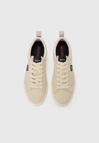Beige sneakers with a textured canvas upper, lace-up design, and branded tongue. Features a loop tab for easy wear, contrasting black insole.