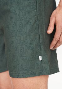 Vihreät kangashousut, joissa on toistuva kuvio viivapiirroksilla olevista hahmoista ja muotokuvista. Mukana on pieni brändilogo sivusaumassa.