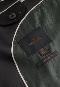 Veste noire avec doublure verte à motifs montrant des étiquettes indiquant "Next Signature," taille 40R, fabriquée au Vietnam, et "Regular Fit."