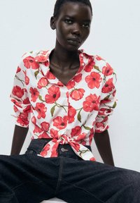 Camicia a fiori con bottoni, con papaveri rossi su sfondo bianco, con maniche arrotolate e un nodo anteriore, abbinata a jeans in denim scuro.