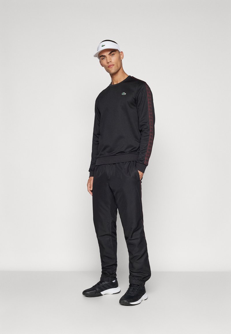 Sweatshirt sportif noir avec des rayures latérales rouges, pantalon noir assorti et baskets noires ; comporte un petit logo sur la poitrine et une casquette à visière.