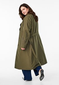 Olijfgroene lange jas met een riem, gemaakt van een licht getextureerde stof en met bloemembroideries op de mouwen; model draagt een breed uitlopende jeans en hoge hakken.