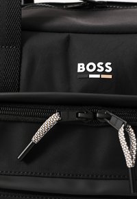 Geantă din țesătură texturată neagră, cu logo-ul "BOSS" în alb, însoțit de dungi negre și bej. Include un fermoar dublu cu mânere modelate.