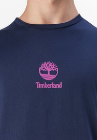 Námořnická modrá bavlněná tričko s klasickým kulatým výstřihem; má růžové kulaté logo a text "Timberland" pod ním. Měkká textura, ležérní design.