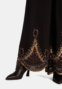Zwarte kledingstuk met ingewikkelde gouden en rode borduursels aan de zoom, met een uitlopende ontwerp. Gecombineerd met een puntige zwarte hoge laars.