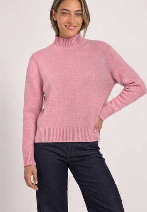Femme souriante portant un pull à col roulé rose et un jean bleu foncé, debout avec une main sur la hanche devant un fond uni.