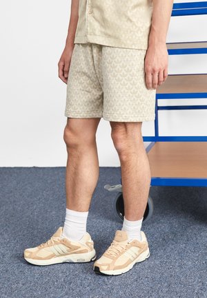 Man die beige, met patroon bedrukte korte broek en bijpassend overhemd draagt, staand op een blauw tapijt bij een blauwe kar, met beige en witte sneakers en witte sokken.