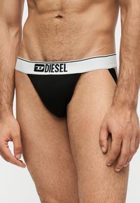 Torse masculin portant un jockstrap noir avec une ceinture blanche portant la marque Diesel, montrant l’abdomen, le haut des jambes et les mains.