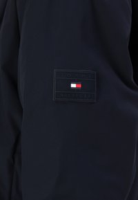 Námořnická bunda s obdélníkovou černou látkovou nášivkou na rukávu s logem "TOMMY HILFIGER" a tri-barvovými akcenty v červené, bílé a modré.