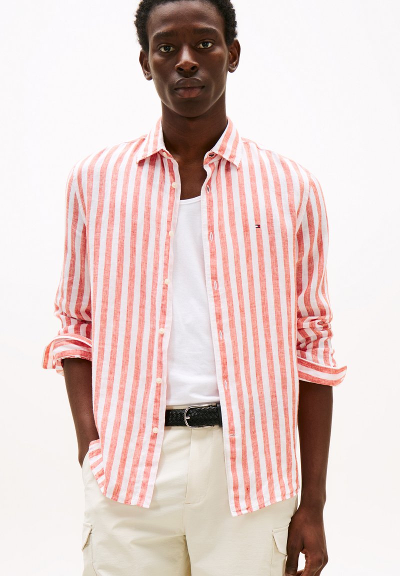 Chemise rayée rouge et blanche, en tissu léger, col boutonné, manches retroussées, portée sur un T-shirt blanc, associée à un pantalon beige.