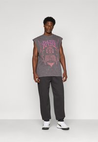 Mand står iført en ærmeløs grå grafisk T-shirt, sorte joggerbukser og hvide sneakers foran en ensfarvet lys baggrund.