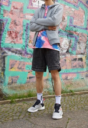Osoba stojąca z założonymi rękami, ubrana w szarą bluzę z kapturem, koszulkę tie-dye, czarne szorty, białe skarpetki i trampki na tle kolorowej ściany z graffiti.