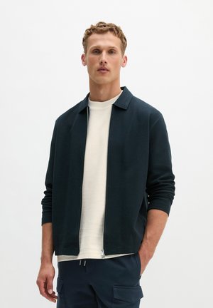 Jonge man die een marineblauw jack met rits draagt over een wit T-shirt met ronde hals, marineblauwe cargobroek, staand tegen een effen witte achtergrond.