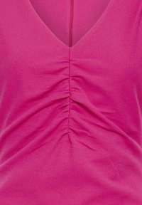 Haut rose à col en V avec un détail froncé rassemblé au centre devant, créant des plis texturés dans le tissu.