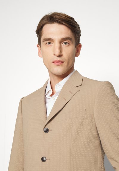 Blazer bege texturizado com lapela em bico, dois botões pretos e um bolso no peito, usado sobre uma camisa branca. Ajuste limpo e sob medida.