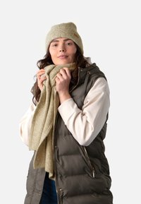 Olijfgroene puffer vest, beige gebreide beanie en zachte sjaal. Model houdt sjaal vast en draagt een licht sweater met manchet. Eenvoudig ontwerp, neutrale tinten.