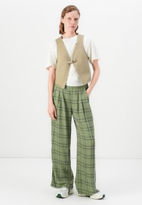 Kvinne med beige vest, hvit t-skjorte, grønne rutete vide bukser og hvite sneakers, stående med en hånd i lommen foran en hvit bakgrunn.