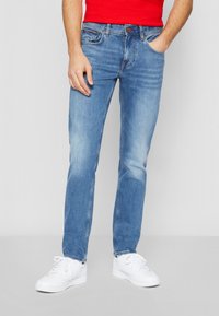 Hellblaue Denim-Jeans mit geradem Schnitt, subtilen Verwaschungen, fünf Taschen und Metallbeschlägen; kombiniert mit weißen Sneakers.