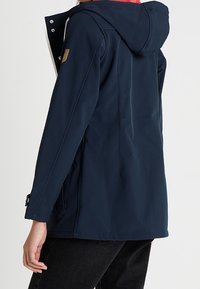 Navy waterproof jacket with a hood, featuring a smooth texture, side pockets, and contrasting inner lining. Gold logo accent on shoulder.