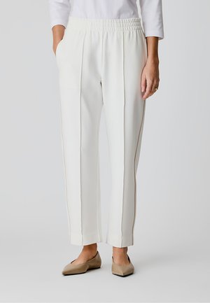 Kvinde iført hvide bukser med lige ben og elastisk talje samt beige spidse sko, stående med den ene hånd i lommen.