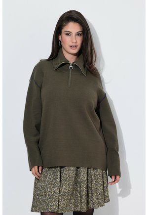 Woman with long dark hair wears olive green zip-up sweater and patterned skirt, standing against a plain light background.