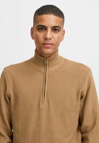 Hellbrauner, zipfeliger Pullover mit geripptem Kragen, strukturiertem Muster und einem viertel-langen Reißverschluss. Er hat ein tailliertes Design und nahtlose Schultern.
