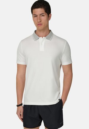 Mężczyzna ubrany w białą koszulkę polo z krótkim rękawem, szarym kołnierzykiem i czarne spodenki, stoi z jedną ręką w kieszeni na jednolitym tle.