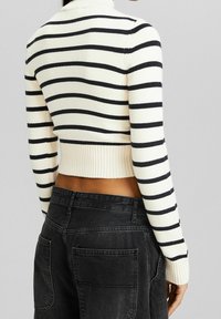 Skuren polotröja med krämfärgade och svarta horisontella ränder, ribbad nederkant, ihopparad med högmidjade svarta jeans, vy från baksidan.