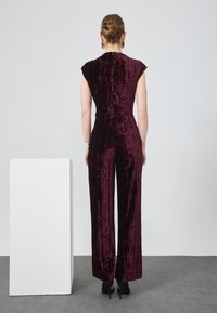 Combinaison en velours bordeaux avec des rayures verticales, des manches courtes et une taille ajustée. Conception à jambes larges associée à des talons hauts noirs.