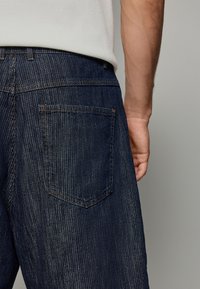 Jeans din denim cu o nuanță albastru închis, prezentând dungi verticale, un singur buzunar pe spate și cusături contrastante. Material texturat vizibil.
