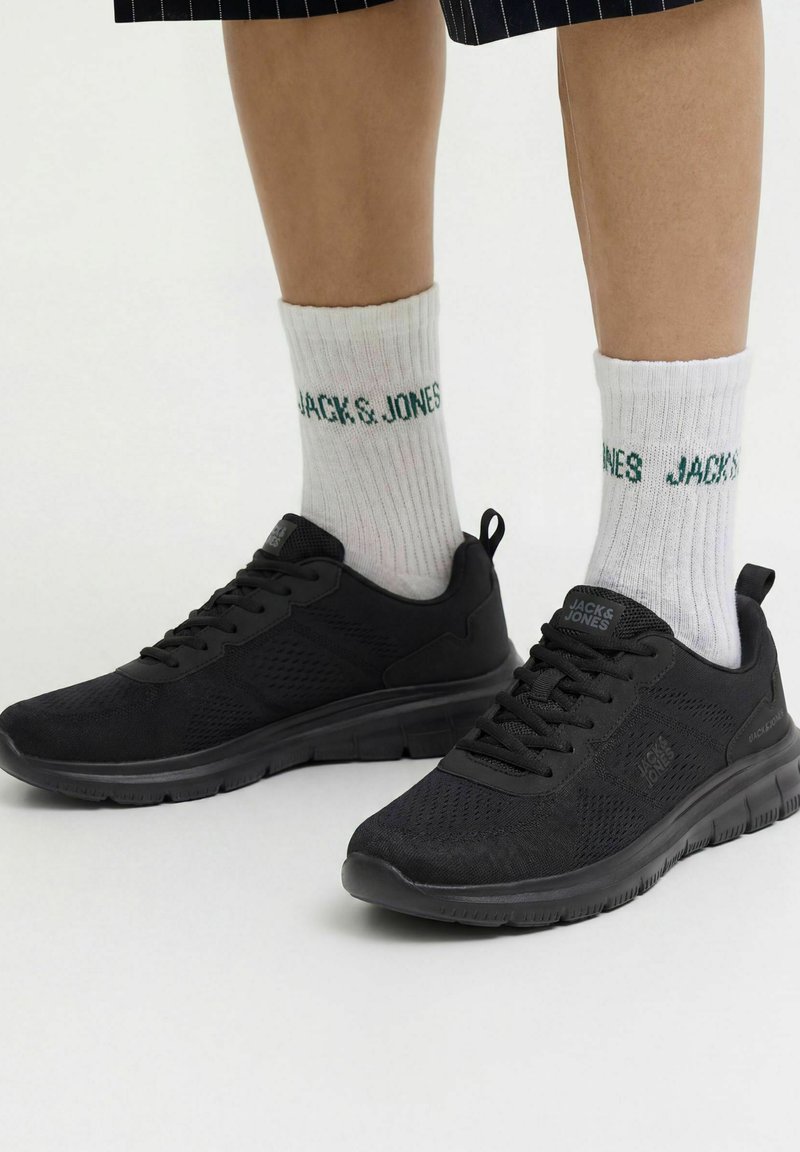 Personne portant des baskets noires Jack & Jones et des chaussettes blanches Jack & Jones, debout sur un fond clair avec une jupe à fines rayures visible.