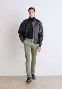 Černá kožená bomber bunda, přiléhavý černý rolák, olivově zelené kalhoty a černé boty. Model stojí před čistým bílým pozadím.