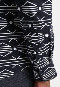 Zwarte shirt met witte geometrische patronen, voorzien van een knoopsluiting bij de manchetten, gemaakt van een gladde stof, die modern ontwerp toont.