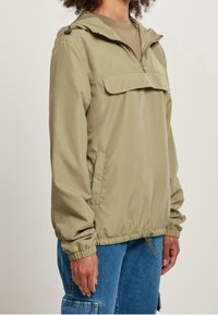 Khaki lekka kurtka z półzamkiem, kieszenią z przodu, regulowanym kapturem, elastycznymi mankietami i ściągaczem u dołu. Gładki, wodoodporny materiał.