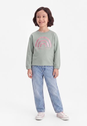 Smilende barn med kort mørkt hår iført en lysegrøn sweatshirt med lyserødt regnbuegrafik, lyseblå jeans og hvide sneakers, stående.