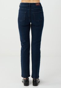 Persona che indossa jeans slim-fit blu scuro e scarpe eleganti nere, in piedi con la schiena rivolta verso la macchina fotografica su uno sfondo semplice.