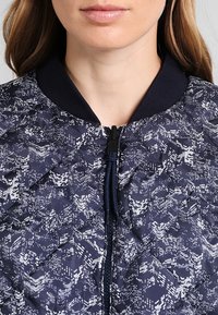 Navyblauer, gesteppten Anorak mit geometrischem weißen Muster, Rippkragen und Frontreißverschluss. Der Stoff hat eine glatte, glänzende Textur.