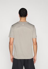 Αθλητική κοντομάνικη μπλούζα σε ανοιχτό γκρι χρώμα, κατασκευασμένη από διαπνέον υλικό, με κοντά μανίκια, στρογγυλή λαιμόκοψη και λεπτομέρεια λογότυπου κοντά στη λαιμόκοψη.