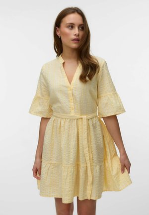 Žena nosi žlté textúrované šaty s volánovými rukávmi do lakťa, výstrihom do V, zapínaním na gombíky vpredu a zaväzovacím opaskom v páse.