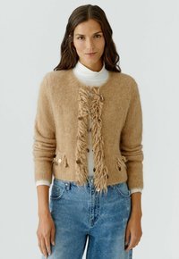 Beige gebreide cardigan met een franjes aan de voorkant, metallic accenten en geribbelde manchetten, gedragen over een witte coltrui en gecombineerd met een blauwe spijkerbroek.