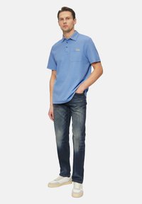 Hellblaues Poloshirt mit einer Tasche, kurzen Ärmeln und drei Knöpfen; kombiniert mit dunklen Jeans und weißen Sneakers mit beigen Akzenten.
