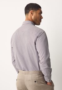 Chemise à manches longues blanche avec un motif bleu, dotée d'un col et d'une coupe ajustée. Portée avec un pantalon ajusté marron clair.