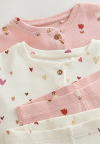 Tre langærmede bluser i pink og hvid vaffelstrik. Mønstrene inkluderer små hjerter og blomster. Hver bluse har en knaplukning ved halsen.