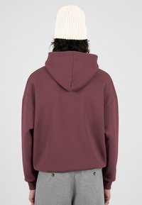 Persona vestida con una sudadera color burdeos, pantalones grises con detalles de botones, y un gorro de punto blanco, vista desde atrás contra un fondo liso.