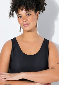 Musta ilmanhiha toppi, jossa on v-pituinen kaula-aukko, sileä rakenne ja muodikas malli. Mallilla on kiharat hiukset ja metalliset pisaramaiset korvakorut.