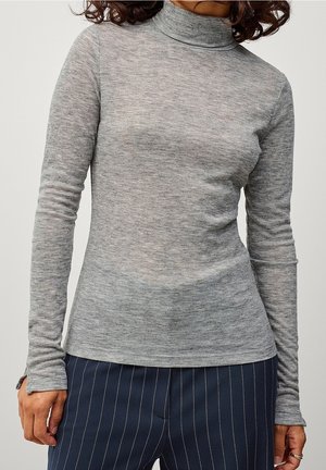 Femme portant un haut ajusté gris clair à col roulé manches longues et un pantalon bleu marine foncé à fines rayures, debout avec les bras détendus le long du corps.